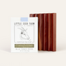 A reddish-brown goat milk soap bar with curved ridges is displayed next to Little Seed Farm’s Soap Bar + Dish Set, featuring a Blue Tansy Rose scent and a handmade cedar dish with a charming goat illustration on the box.