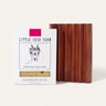 A Little Seed Farm Soap Bar + Dish Set features a milk soap bar in white packaging with goat illustrations next to a brown, ridged handmade cedar soap dish.