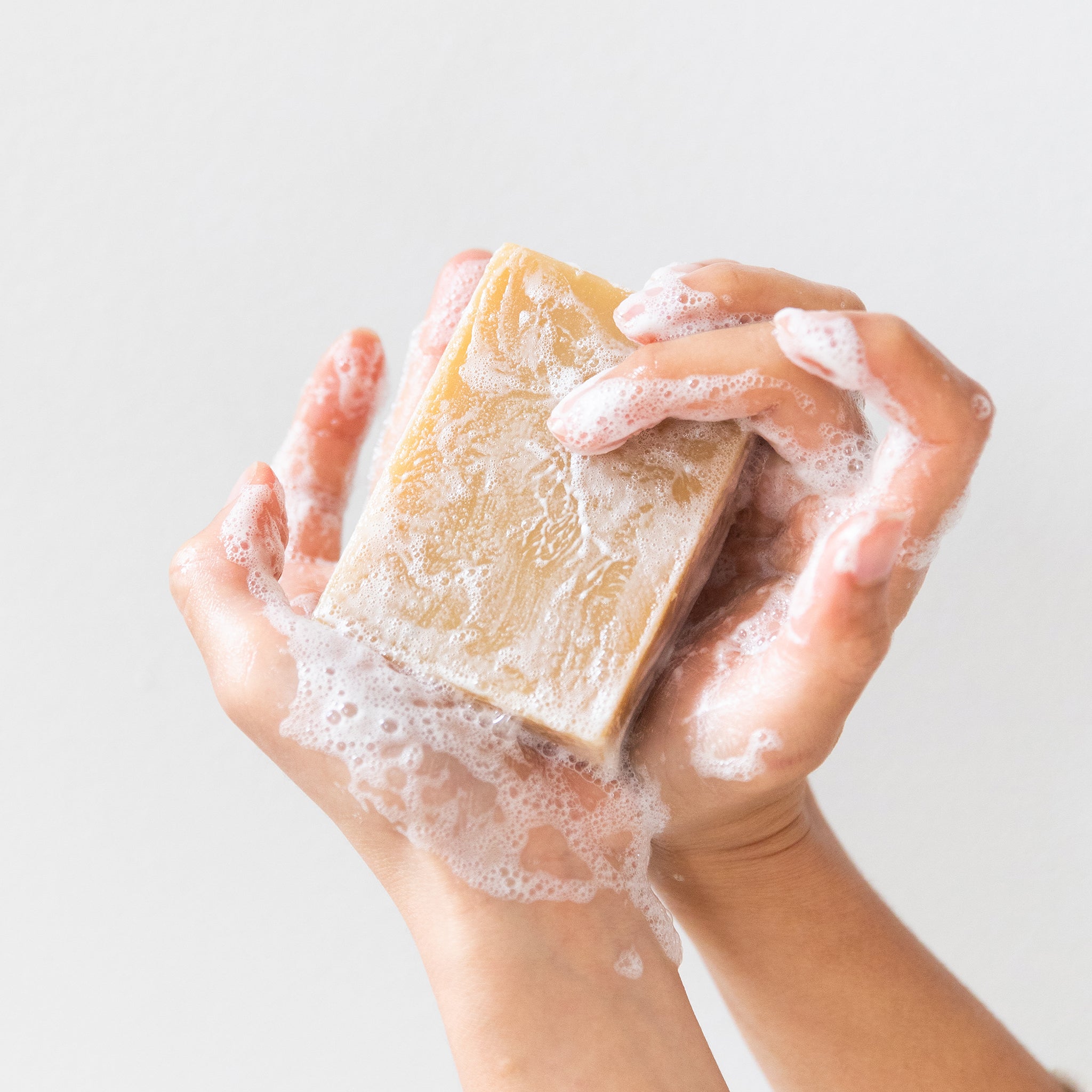 Two hands hold a large Little Seed Farm Soap Bar + Dish Set, the soap covered in white, foamy lather against a plain, light background.