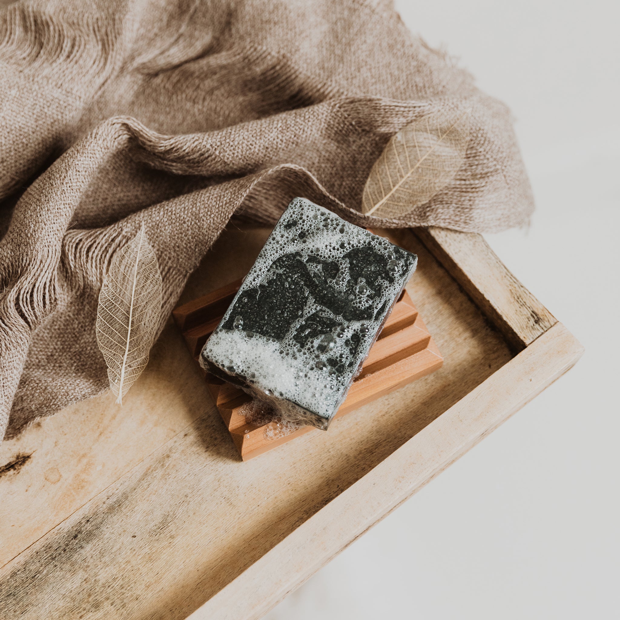 A Soap Bar + Dish Set from Little Seed Farm sits on a rustic tray with a textured brown cloth and two skeleton leaves beside it.