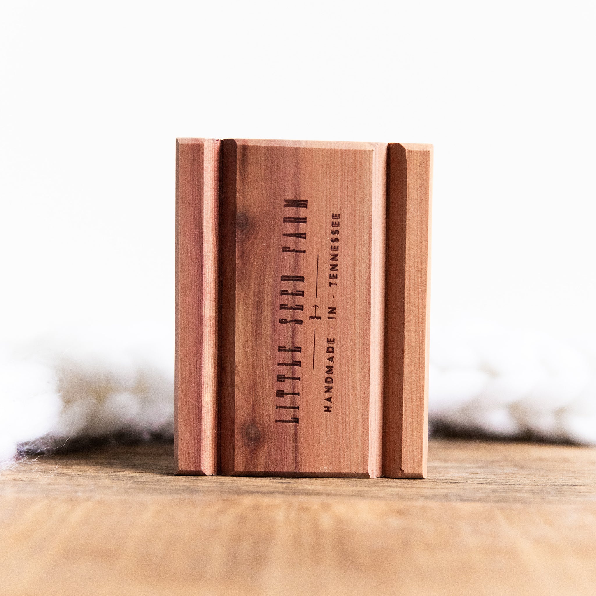 A rectangular wooden block stamped with Little Seed Farm stands upright beside a Soap Bar + Dish Set, displayed on a wooden surface with soft white material blurred in the background.