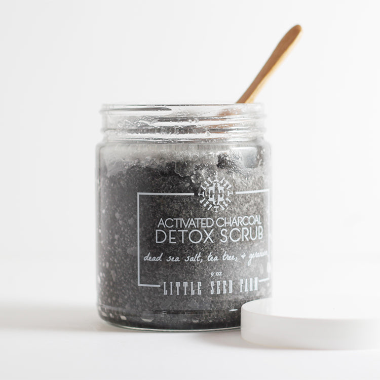 Activated Charcoal Detox Scrub in a refillable glass jar with a bamboo scoop, featuring a deep-black exfoliant made with Dead Sea salt, activated charcoal, and organic coconut oil to detoxify and moisturize.