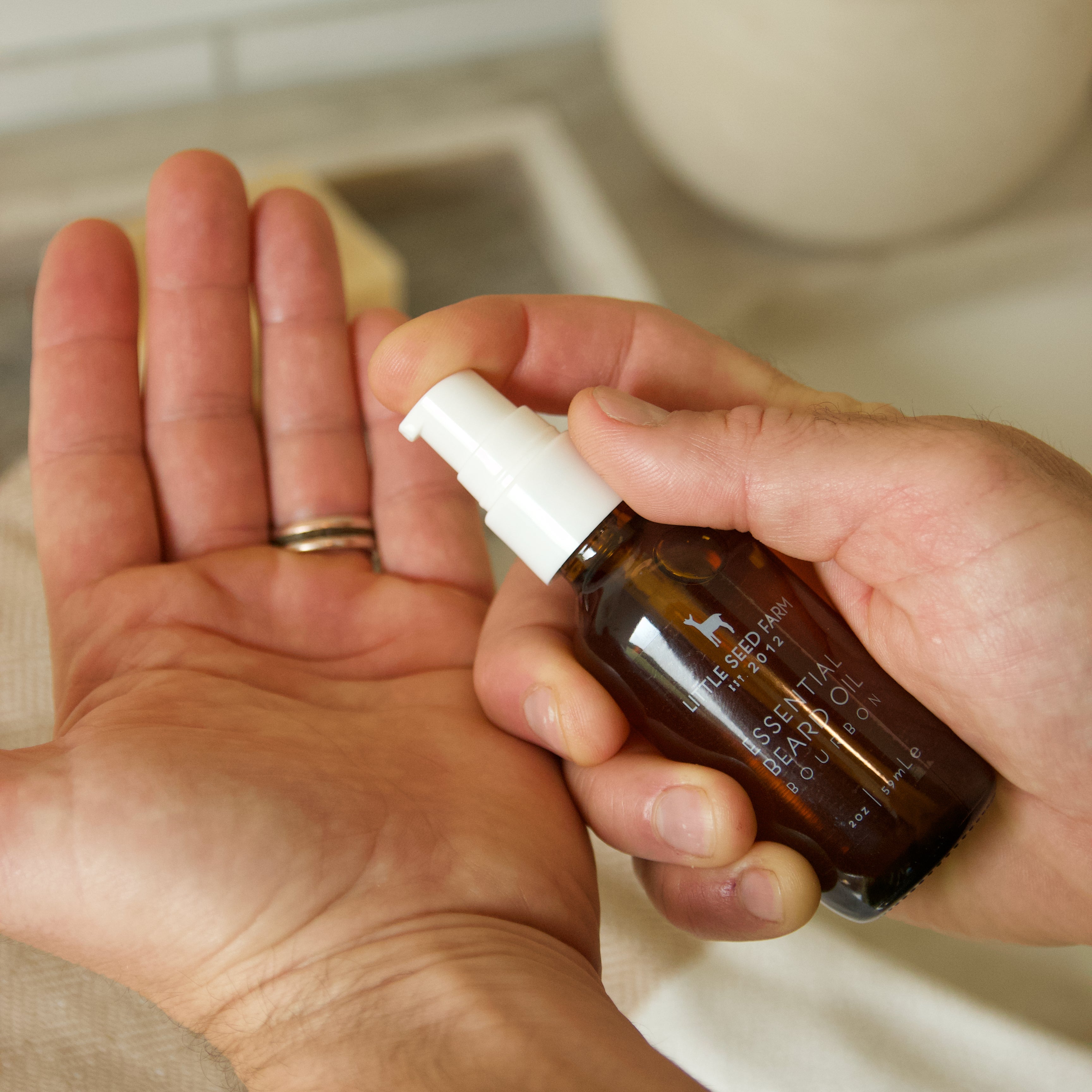 Person holding a bottle of beard oil over their palm, with a plant in the background.